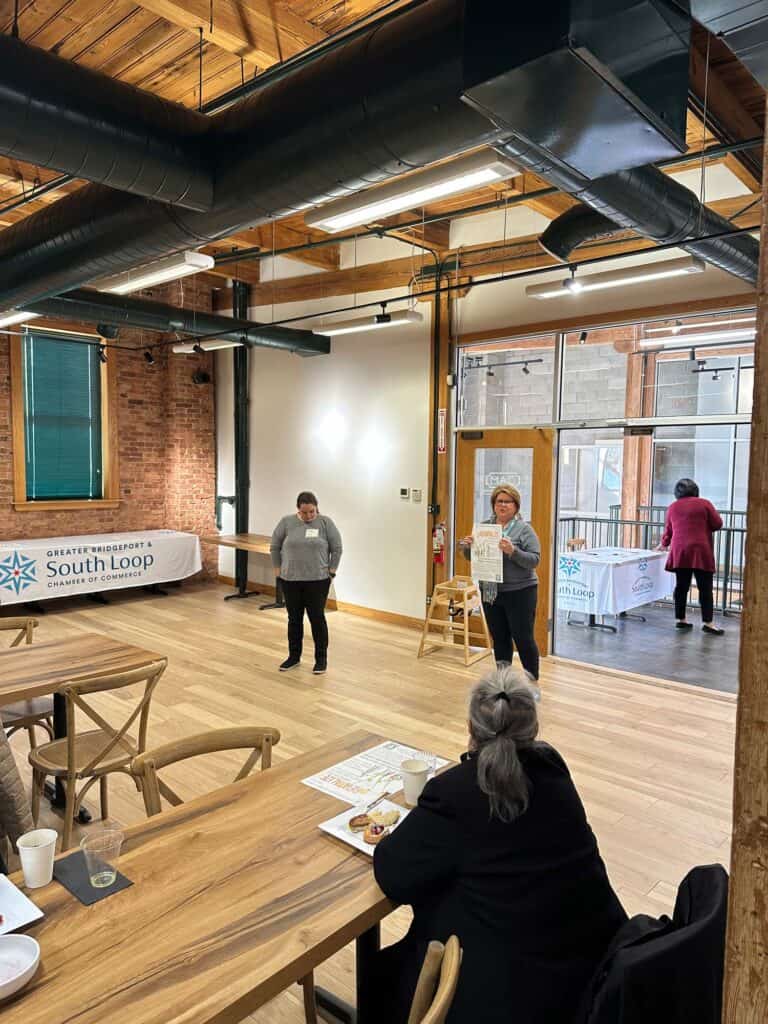 A speaker addresses a small audience in a spacious room with wooden floors and exposed beams. Two banners reading South Loop Chamber of Commerce are displayed. Attendees are seated at a wooden table with drinks and papers.