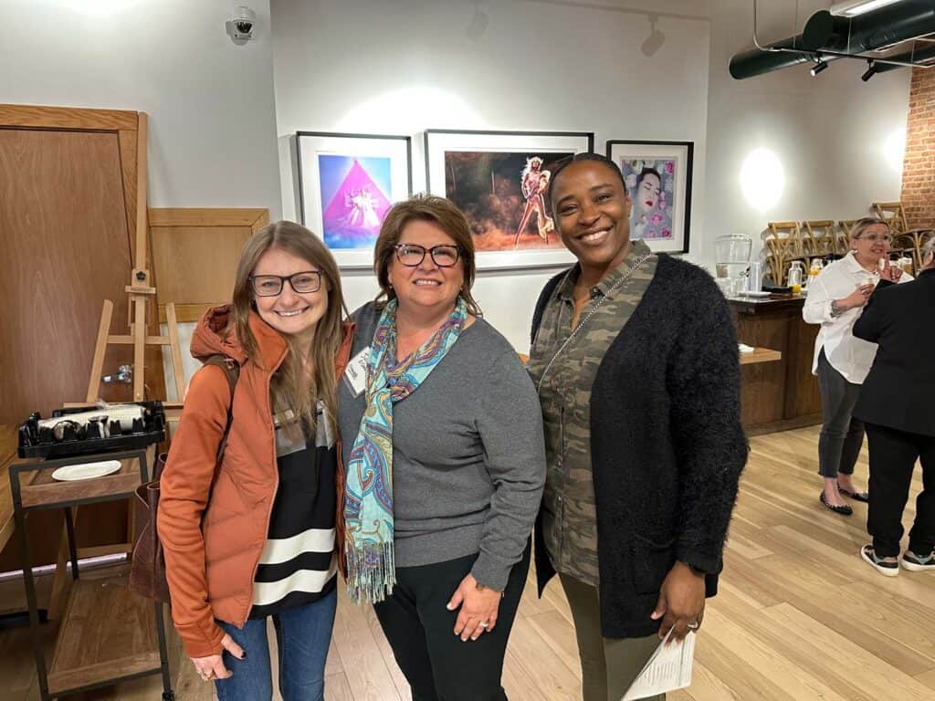 Three people stand smiling in an art gallery. The first person wears glasses and a red jacket, the second person wears glasses and a scarf, and the third person wears a black cardigan. Artworks hang on the wall behind them.