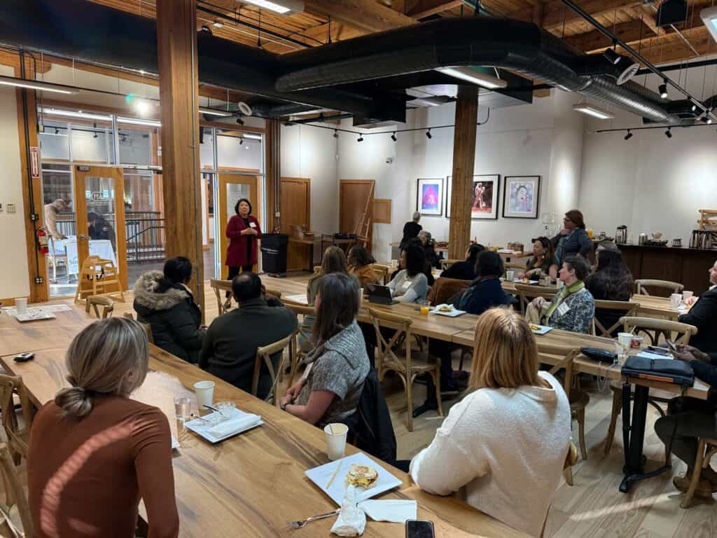 A woman stands speaking to a group of people seated at long wooden tables in a well-lit room with artwork on the walls. The location appears to be a café or meeting space with high ceilings and modern decor.