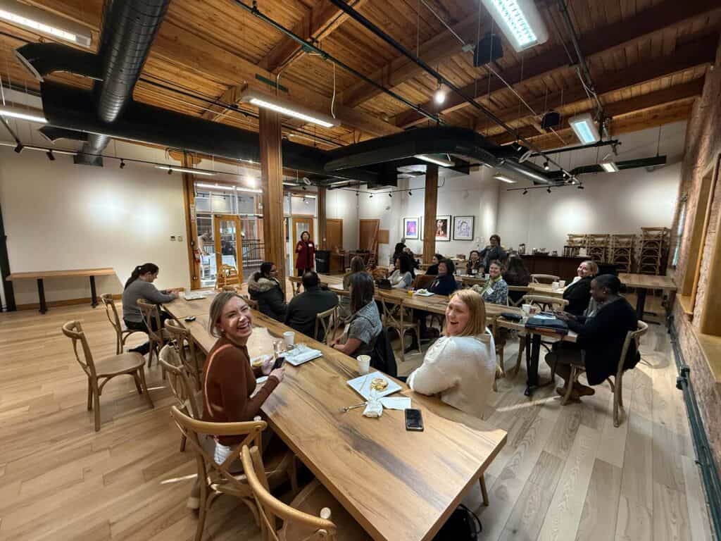 A group of people seated around wooden tables in a large, well-lit room with a wooden ceiling and beams. Some are engaged in conversation, while others are focused on their meals. A few are smiling towards the camera.
