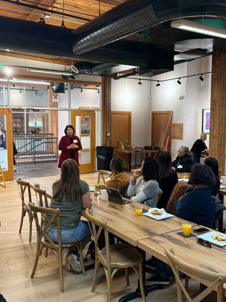 A person is standing and speaking to a seated audience in a modern conference room. The room features wooden decor and a long table with plates and drinks. Attendees are listening attentively.
