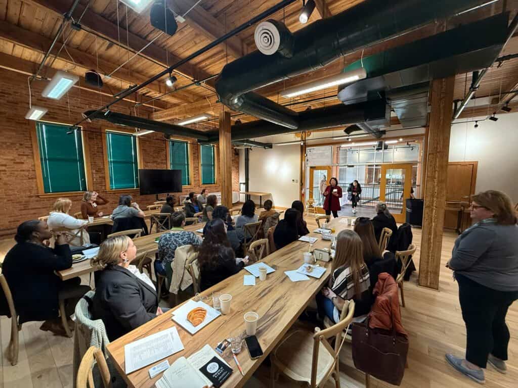 A group of people seated at long wooden tables attentively listening to two presenters at the front of an industrial-style room. The room has exposed brick walls, beams, and large windows. Various notebooks and drinks are on the tables.