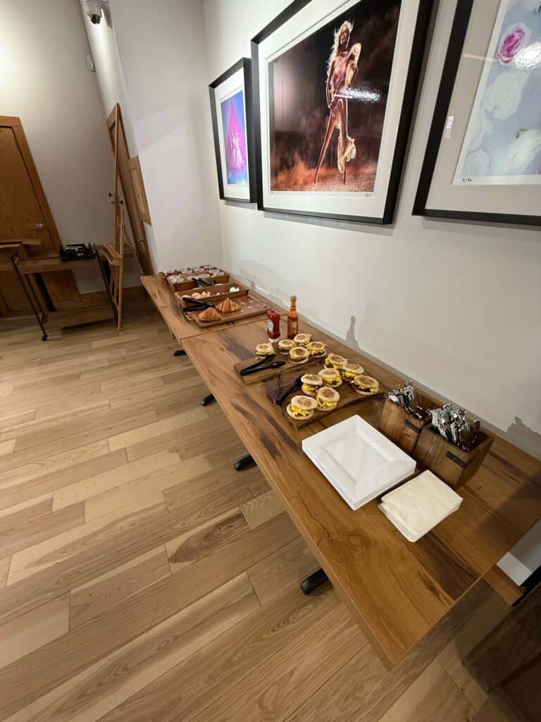 A wooden table set with pastries, toasted bread, bottles of hot sauce, cutlery, and napkins. Framed photos hang on the wall behind, and an easel is visible in the corner of the room with wooden flooring.