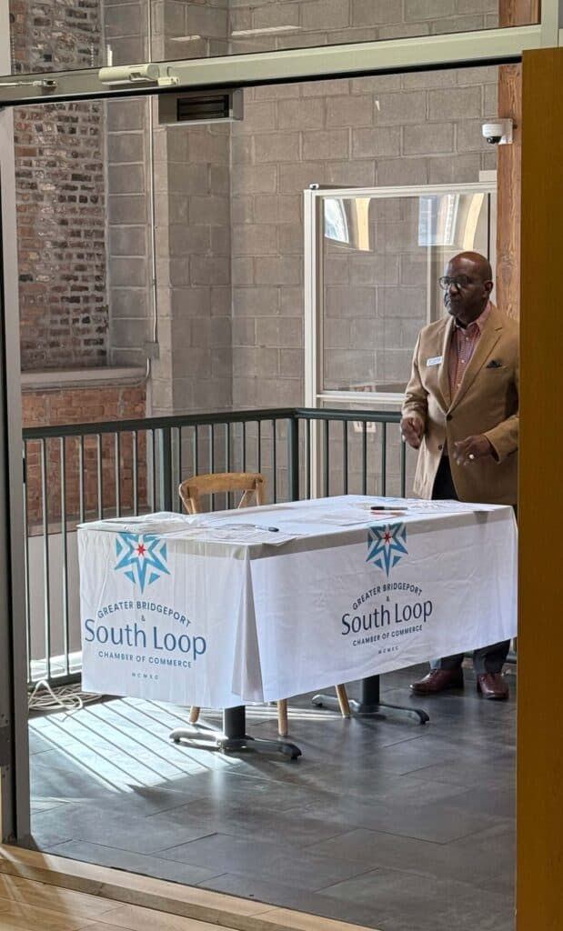 A man stands behind a table covered with a cloth labeled Greater Bridgeport, South Loop, Chamber of Commerce. He appears to be speaking or presenting. The setting is indoors, with brick walls and windows in the background.