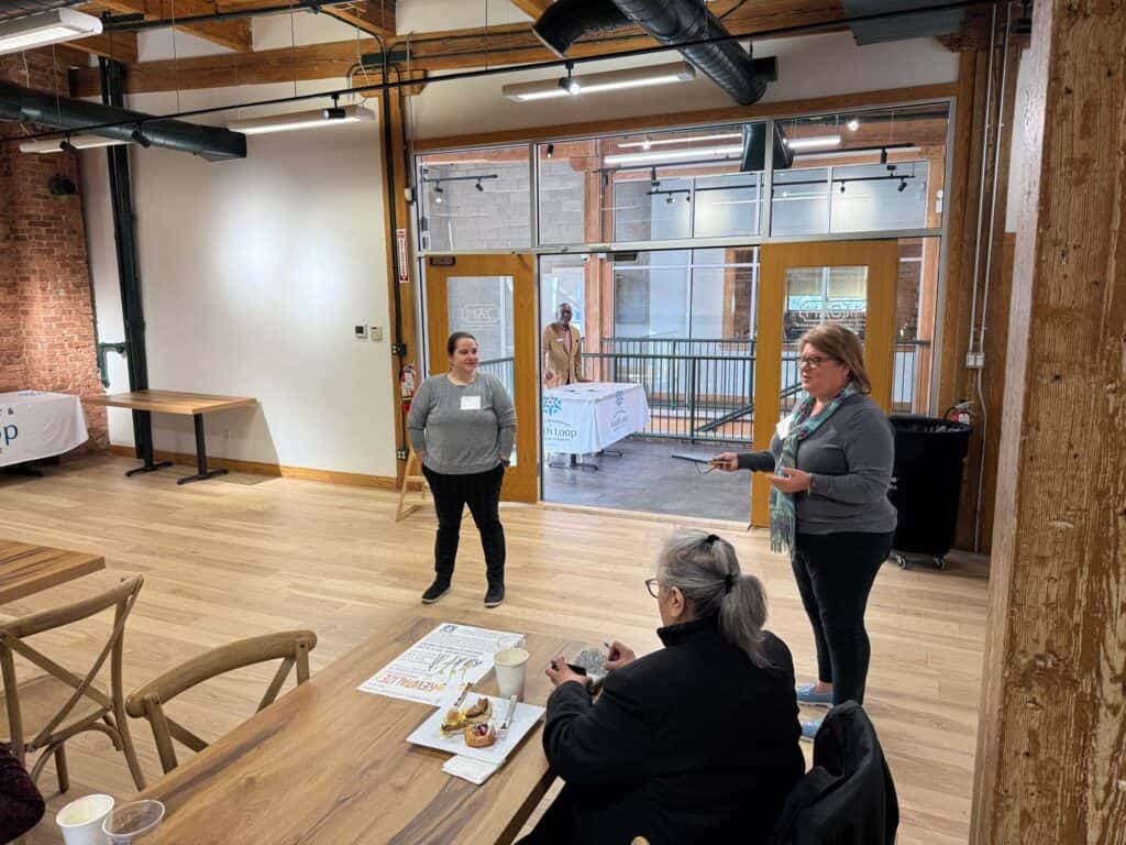 Two women are standing and speaking in a room with wooden floors and exposed brick walls. One holds a microphone. Three people are seated at a table with drinks. A large window shows a person outside at a table with a banner.
