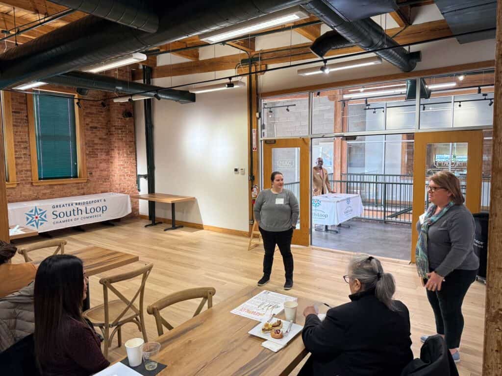 Three people are gathered in a modern office space with brick walls and wooden floors. Two individuals are standing and talking to a seated person. A South Loop Chamber of Commerce banner is visible. Natural light streams in through large windows.