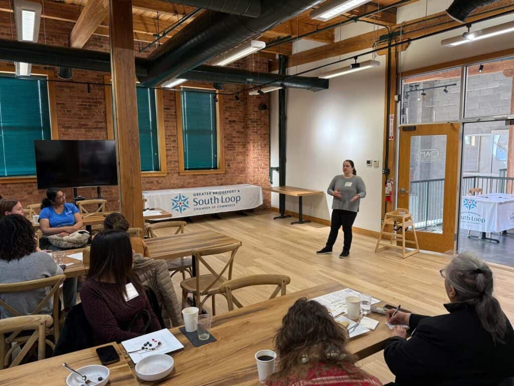 A woman stands speaking in front of a small audience seated at tables in a bright room with brick walls and large windows. Theres a banner reading South Loop and a TV on the wall. Coffee cups and papers are on the tables.