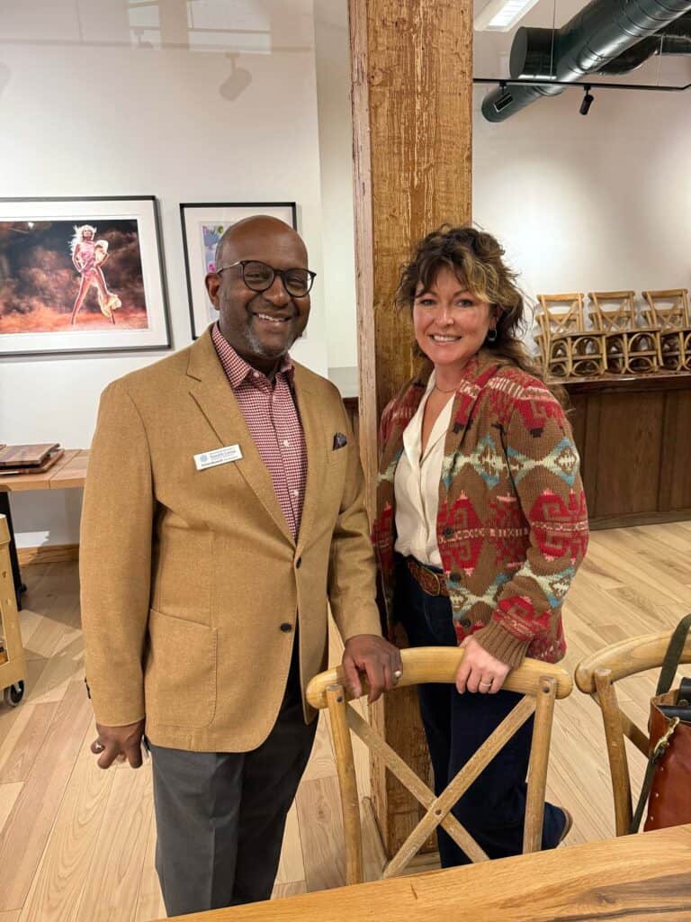 Two people smiling while standing indoors near wooden chairs. The man wears a tan blazer, glasses, and has a name tag. The woman wears a patterned cardigan. Theres wood flooring, exposed beams, and framed art on the wall in the background.