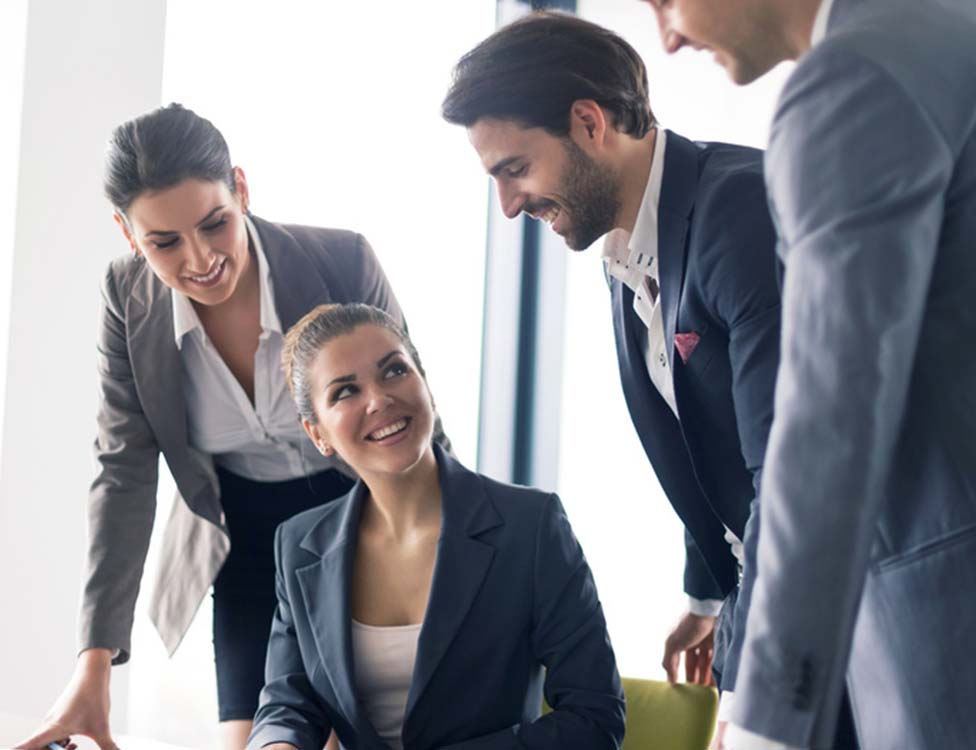 Four professionals in business attire are gathered around a table, engaged in a positive discussion. Two women and two men are smiling, suggesting a collaborative and friendly atmosphere in a bright office setting.